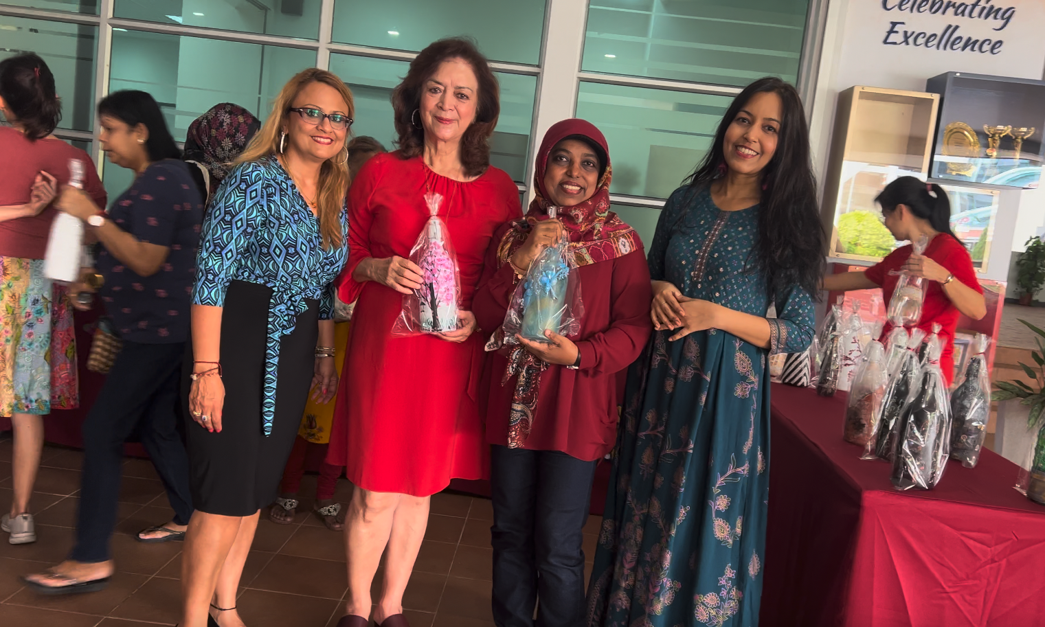 Four women smile together at a Celebrating Excellence event, two holding wrapped decorative bottles as gifts.